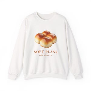May include: A white long-sleeve crewneck sweatshirt featuring a graphic of four golden-brown dinner rolls centered on the chest. Below the rolls, the text reads "SOFT PLANS" in large letters and "soft plans co." in smaller text below it.