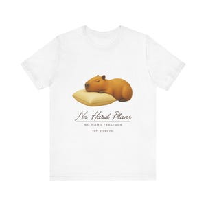 May include: A white short-sleeve t-shirt featuring a graphic of a brown capybara sleeping on a tan pillow. Below the illustration, the text reads "No Hard Plans," "NO HARD FEELINGS," and "soft plans co." in black, stylized, and sans-serif fonts.