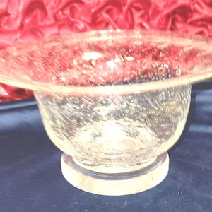 May include: A clear glass bowl with a textured surface. The bowl has a wide, shallow rim and sits on a small, circular base. The glass appears to have a pattern of small bubbles or textures throughout.