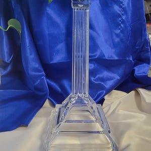 May include: A clear, crystal candlestick holder with a tall, fluted column and a square base. The top features a square cup to hold a candle. The background is a royal blue fabric.