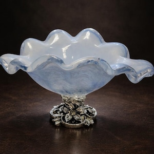 May include: A decorative glass bowl with a ruffled rim and a silver-colored floral base. The bowl has a light blue and white color scheme, with a wavy, cloud-like design. The bowl is a decorative item.