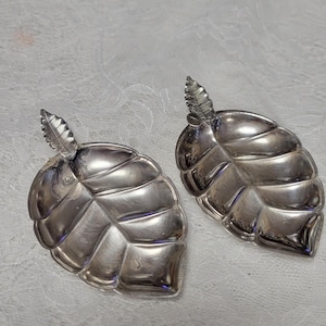 May include: Two silver-colored leaf-shaped dishes. Each dish has a detailed leaf vein pattern and a small, textured leaf at the top. The dishes are displayed on a white, patterned surface.