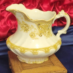 Pode incluir: Uma jarra de cerâmica branca vintage com padrões florais e acabamentos amarelos. A jarra tem uma alça curva e uma borda ondulada. É exibida em um pequeno suporte de madeira esculpida.
