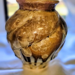 Może przedstawiać: Ręcznie wykonany ceramiczny wazon o unikalnym, organicznym kształcie. Wazon ma brązową podstawę z wirującymi wzorami i białymi akcentami, zwieńczoną czerwonobrązową obwódką. Błyszczące wykończenie podkreśla szczegółową fakturę.