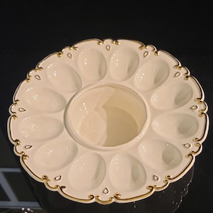 May include: A round white ceramic serving platter featuring twelve oval-shaped indentations arranged in a circle around a central bowl. The scalloped edge of the platter is decorated with a thin gold trim and small gold accents between each section.