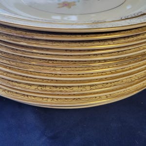 Könnte beinhalten: Ein Stapel cremefarbener Vintage-Teller mit goldenen Verzierungen und floralen Mittelmuster. Die Teller haben ein zartes, verziertes Goldmuster an den Rändern. Das florale Design umfasst rosa, blaue und gelbe Blumen.