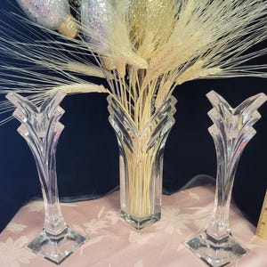 May include: A clear glass vase and two matching candle holders. The vase holds a bouquet of wheat and glittery gold and silver ornaments. The items are on a pink floral patterned surface.