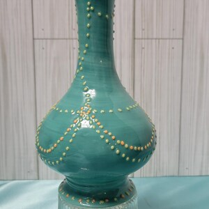 K&ouml;nnte beinhalten: Eine t&uuml;rkisfarbene Keramikvase mit bauchigem K&ouml;rper und einem Sockel aus klarem Glas. Die Vase ist mit orangefarbenen und gelben Punktmustern verziert. Die Vase hat eine gl&auml;nzende Oberfl&auml;che und wird vor einem hellen Holzpaneel-Hintergrund pr&auml;sentiert.