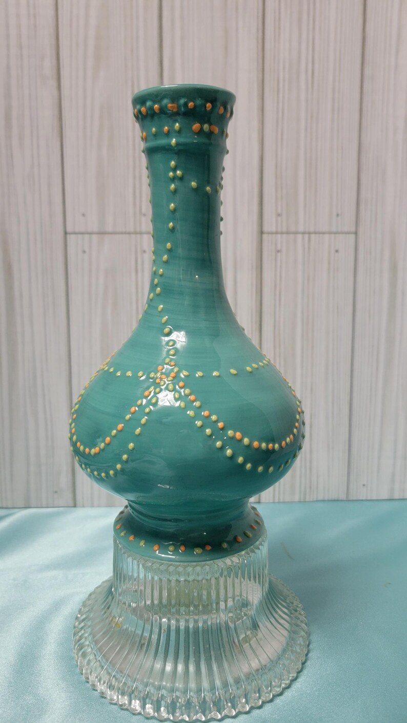 K&ouml;nnte beinhalten: Eine t&uuml;rkisfarbene Keramikvase mit bauchigem K&ouml;rper und einem Sockel aus klarem Glas. Die Vase ist mit orangefarbenen und gelben Punktmustern verziert. Die Vase hat eine gl&auml;nzende Oberfl&auml;che und wird vor einem hellen Holzpaneel-Hintergrund pr&auml;sentiert.