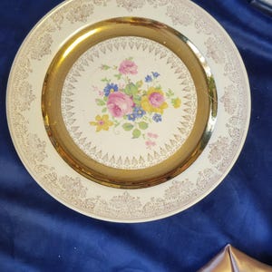 Könnte beinhalten: Vintage Zierteller mit floralem Design. Der Teller hat einen goldenen Rand und eine zentrale Blumenanordnung aus rosa, blauen und gelben Blumen. Der äußere Rand hat einen cremefarbenen Hintergrund mit einem goldenen Filigranmuster.
