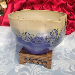 May include: A handcrafted ceramic bowl with a unique design. The bowl features a two-tone color scheme, with a beige upper section and a deep blue lower section. The blue section has a textured, speckled appearance. The bowl sits on a decorative wooden stand.