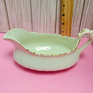 May include: Cream-colored gravy boat with a scalloped rim and a gold trim. The gravy boat has a handle and a spout for easy pouring. The interior is a light cream color. A wooden ruler is in the background.