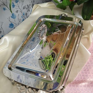 May include: A rectangular, silver-colored serving tray with ornate detailing. The tray has a highly reflective surface, with decorative handles and feet. A small, decorative element is attached to the tray's surface. The tray is likely used for serving food or displaying items.