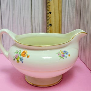 May include: Cream-colored ceramic gravy boat with a handle and spout, adorned with floral patterns in shades of orange, blue, and pink. The rim and base are trimmed with gold. A vintage serving dish.