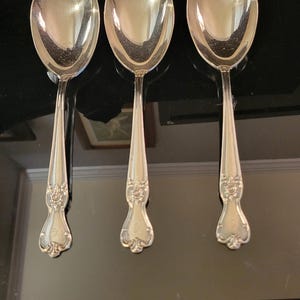 May include: Three silver-toned metal spoons with wide, rounded bowls and slender handles. Each handle features an ornate, embossed floral pattern near the base, which includes a small, engraved letter G centered within the decorative design.