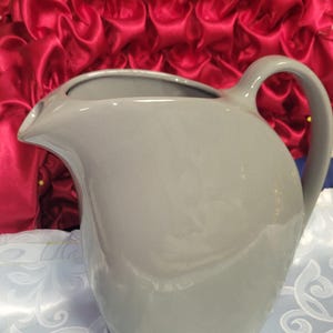 May include: A gray ceramic pitcher with a curved spout and a rounded handle. The pitcher is sitting on a light blue tablecloth with a white floral pattern. A red, ruffled fabric is in the background.