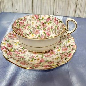 Könnte beinhalten: Ein Vintage-Tee-Set mit Tasse und Untertasse. Die weiße Porzellantasse und Untertasse sind mit einem zarten Blumenmuster in Rosa, Gelb und Grün verziert. Goldene Verzierungen akzentuieren die Ränder.
