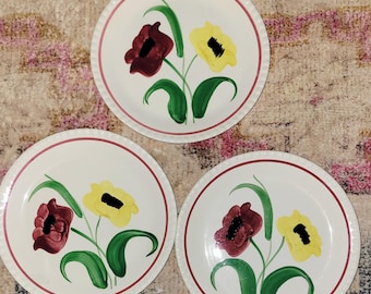May include: Three white ceramic plates with a red rim and a floral design. Each plate features a bouquet of flowers with burgundy and yellow blooms, green leaves, and black centers. The plates are arranged on a patterned surface.