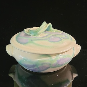 May include: A round ceramic lidded bowl featuring a glossy off-white base glaze with swirling watercolor-style patterns in shades of soft lavender and mint green. The bowl includes two side handles and a sculptural, arched handle on the lid.