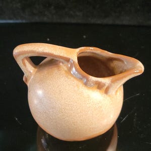 May include: A small, round ceramic creamer with a light tan, speckled glaze and a smooth, glossy finish. It features a single arched handle on one side and a small, pointed pouring spout on the opposite side, with a darker brown rim accent.