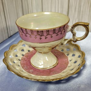 May include: A vintage teacup and saucer set. The teacup has a pink band with gold accents and a gold handle. The saucer is white with a pink center and a gold rim. The set is made of porcelain.