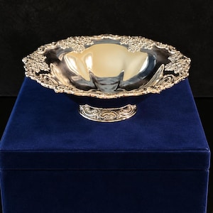 Puede incluir: Un cuenco de pedestal en tono plateado pulido con un borde ornamentado en relieve que presenta detalles intrincados de flores y racimos de uvas. El cuenco tiene una cuenca interior lisa y reflectante y descansa sobre una base circular decorativa.