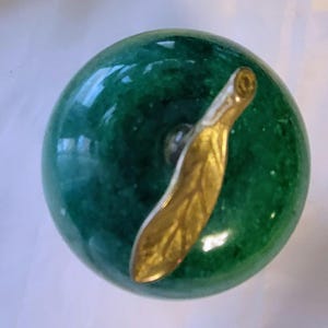 May include: A polished green stone apple figurine with a smooth, speckled texture. The apple features a metallic gold-toned stem and a single leaf attached to the top, creating a decorative piece with a glossy, reflective surface finish.