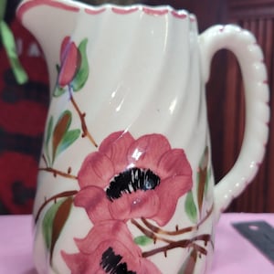 May include: White ceramic jug with a twisted body and a handle. The jug is decorated with large pink flowers with black centres and green leaves. The rim and handle are trimmed in pink.