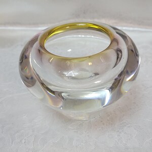 May include: A clear glass bowl with a thick, rounded rim featuring a subtle yellow-green tint. The bowl has a smooth, polished surface and a heavy, solid base, creating a rounded, organic shape that reflects light through its transparent walls.