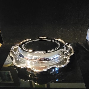 May include: A silver-colored oval serving tray with a polished, reflective surface and a decorative scalloped edge. The tray features two small handles on opposite sides and a beaded border detail surrounding the central flat area of the platter.
