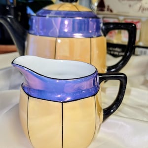 May include: A vintage tea set featuring a teapot and creamer. The set has a cream-colored body with black lines and a blue iridescent band. The teapot has a black handle and spout, and the creamer has a white interior.