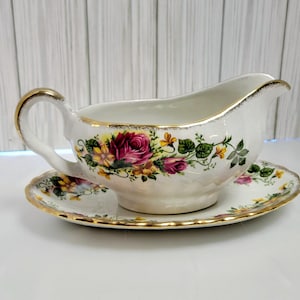 Puede incluir: Salsera de cerámica blanca con platillo a juego. El conjunto presenta un diseño floral con rosas rosas y amarillas, hojas verdes y ribete dorado. La salsera tiene un asa y un pico vertedor.