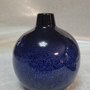 May include: A round, dark blue vase with a speckled texture and a glossy finish. The vase has a narrow, black neck and a spherical body. This decorative vase is suitable for home decor.