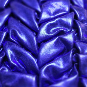 Puede incluir: Primer plano de una tela azul oscuro texturizada y brillante. La tela está doblada y recogida, creando un patrón de formas de diamante superpuestas. El material tiene un ligero brillo, reflejando la luz.