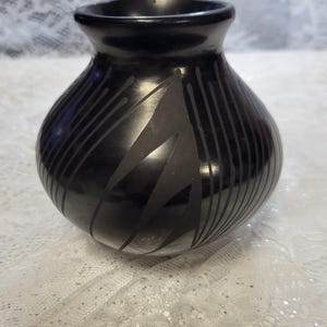 May include: A black ceramic vase with a rounded body and a narrow neck. The vase features a geometric design of angled lines and shapes. The vase is displayed on a white lace surface.