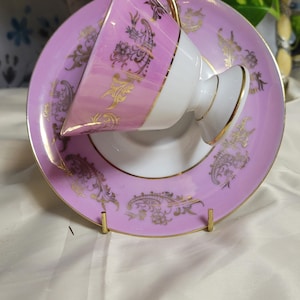 May include: A vintage teacup and saucer set. The cup and saucer are a soft pink with gold filigree designs. The cup has a white interior and a gold handle. The saucer has a matching design. The set is displayed on a gold stand.
