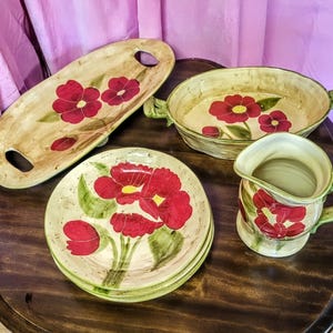 Puede incluir: Un conjunto de fuentes de cerámica pintadas a mano con un diseño floral. El juego incluye una fuente alargada, una fuente para hornear, platos y una jarra. Las flores son rojas con centros amarillos y hojas verdes, sobre un fondo crema.