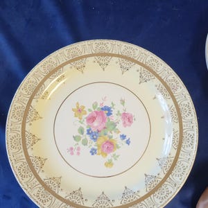 Puede incluir: Un plato vintage de color crema con un diseño floral. El centro presenta flores rosas, amarillas y azules. El borde tiene un patrón de filigrana dorada. Una pieza clásica para servir o exhibir.