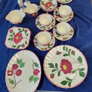 May include: A collection of vintage ceramic tableware with a floral design. The set includes plates, saucers, cups, a creamer, a sugar bowl, and decorative figurines. The floral pattern features red flowers with green leaves and pink trim.