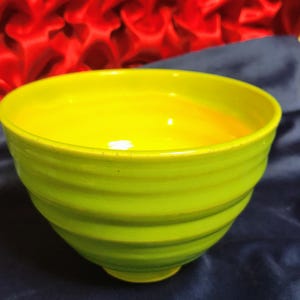 Könnte beinhalten: Eine handgefertigte Keramikschale mit leuchtend gelbem Innenraum und einer strukturierten, limettengrünen Außenseite. Die Schale hat eine abgerundete Form mit leicht ausgestelltem Rand und einem kleinen, kreisförmigen Boden. Die Außenseite weist horizontale, geriffelte Details auf.