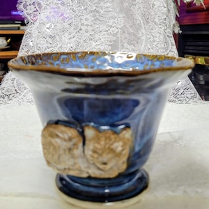 Pode incluir: Uma tigela de cerâmica com borda alargada, apresentando um esmalte azul profundo com detalhes em marrom. O exterior é decorado com um relevo esculpido em marrom claro de um animal enrolado. A borda interna tem um padrão texturizado.