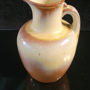 May include: A ceramic pitcher featuring a rounded body and a curved handle. The exterior has a smooth, glazed finish with a gradient color scheme transitioning from a warm, earthy brown at the base to a lighter, creamy beige tone near the top rim.