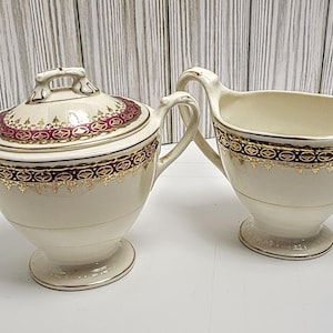 May include: A cream-coloured sugar bowl with lid and a creamer. Both items have a gold and burgundy geometric pattern around the rims and lids. The sugar bowl has a handle on the lid and the creamer has a curved handle.