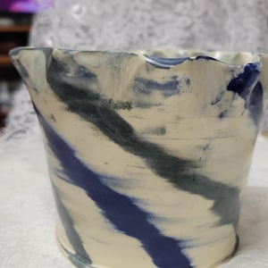 Peut inclure: Un pot en céramique avec un bord évasé et ondulé présentant un motif marbré de stries crème, bleu foncé et gris anthracite. Le récipient a un corps cylindrique qui se rétrécit légèrement vers la base, avec une finition lisse façonnée à la main.