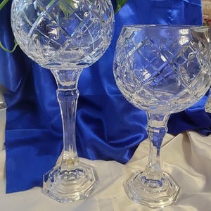 May include: Two clear crystal goblets with a diamond pattern. The goblets have a round bowl, a long stem, and a hexagonal base. They are set against a blue satin background.
