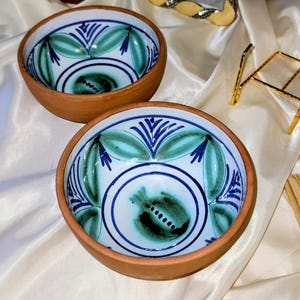 May include: Two terracotta bowls with hand-painted designs. The bowls feature a white interior with blue and teal leaf and floral patterns. The rims are a natural terracotta color.