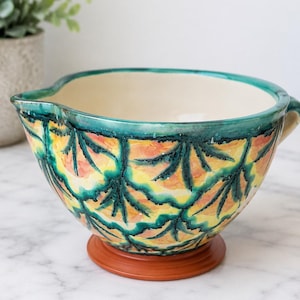 Pode incluir: Uma tigela de mistura em cerâmica com interior creme e um exterior vibrante com um padrão pintado à mão de formas de folhas em azul-petróleo escuro sobre um fundo estilo aguarela amarelo e laranja, assente numa base cor de terracota.