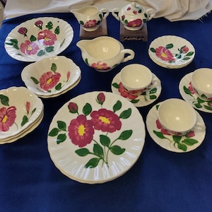 May include: A collection of vintage floral-decorated dishware. The set includes plates, bowls, saucers, and teacups, all featuring a hand-painted design of pink flowers with green leaves. The pieces are arranged on a blue surface.