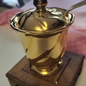 Puede incluir: Un azucarero dorado y ornamentado con tapa y cuchara, sobre un soporte de madera oscura. El cuenco tiene un diseño acanalado y un adorno decorativo en la tapa. La cuchara también es dorada.