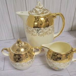 May include: A vintage cream-coloured tea set with gold accents. The set includes a teapot with a lid, a sugar bowl with a lid, and a creamer. The gold detailing features floral and ornate patterns, adding a touch of elegance to the set.